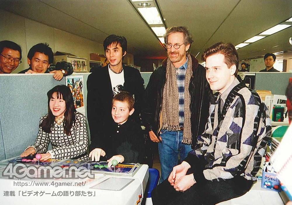 Grupo de pessoas em frente a um monitor, entre elas o diretor de cinema Steven Spielberg e o produtor de games Yu Suzuki