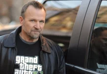 Ator Ned Luke ao lado de um carro, usando uma jaqueta preta e camiseta com logotipo do jogo Grand Theft Auto 5