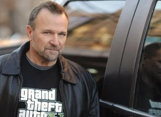 Biografia: Ned Luke Ator Ned Luke ao lado de um carro, usando uma jaqueta preta e camiseta com logotipo do jogo Grand Theft Auto 5