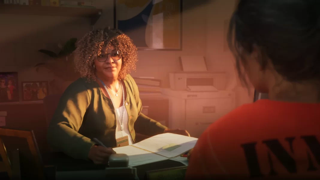 Uma mulher negra de aproximadamente 50 anos, com cabelos longos, sentada em sua mesa de trabalho, conversa com Lucia, sentada à sua frente. Ela parece ser uma funcionária do presídio.