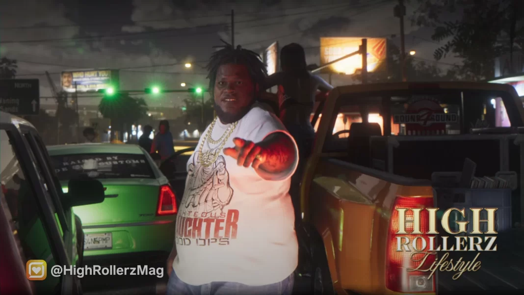 Um homem negro obeso usando camiseta branca aponta para a câmera; há carros ao redor no que parece ser a largada de uma corrida de rua, à noite.